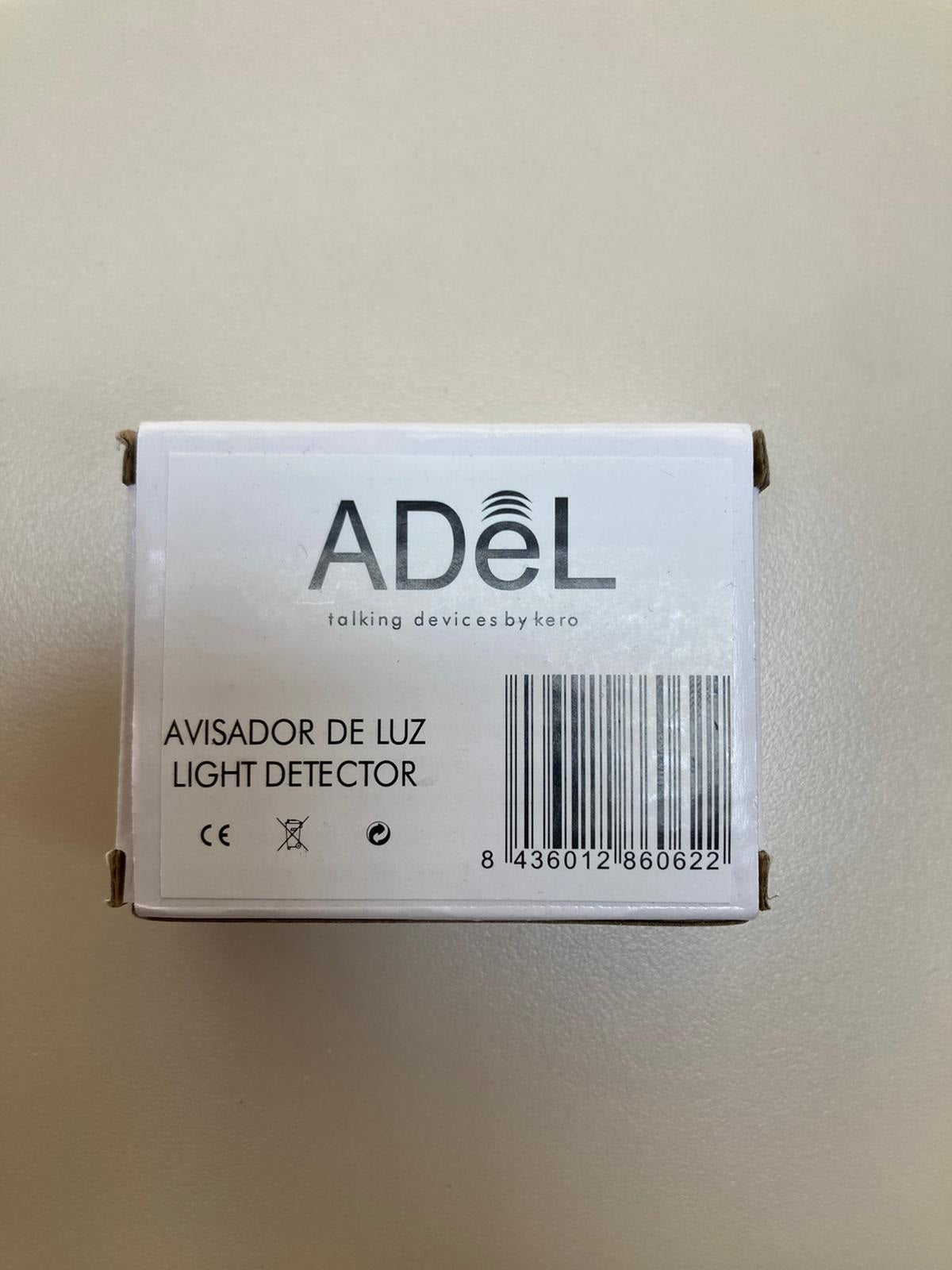 A small white box labeled "Adel Portable Light Detector" is shown on a light-colored surface. The box has the brand name "ADEL" printed prominently on the top, along with the phrase "talking devices by koro." Below that, it says "AVISADOR DE LUZ LIGHT DETECTOR" and includes a barcode and some certification symbols.