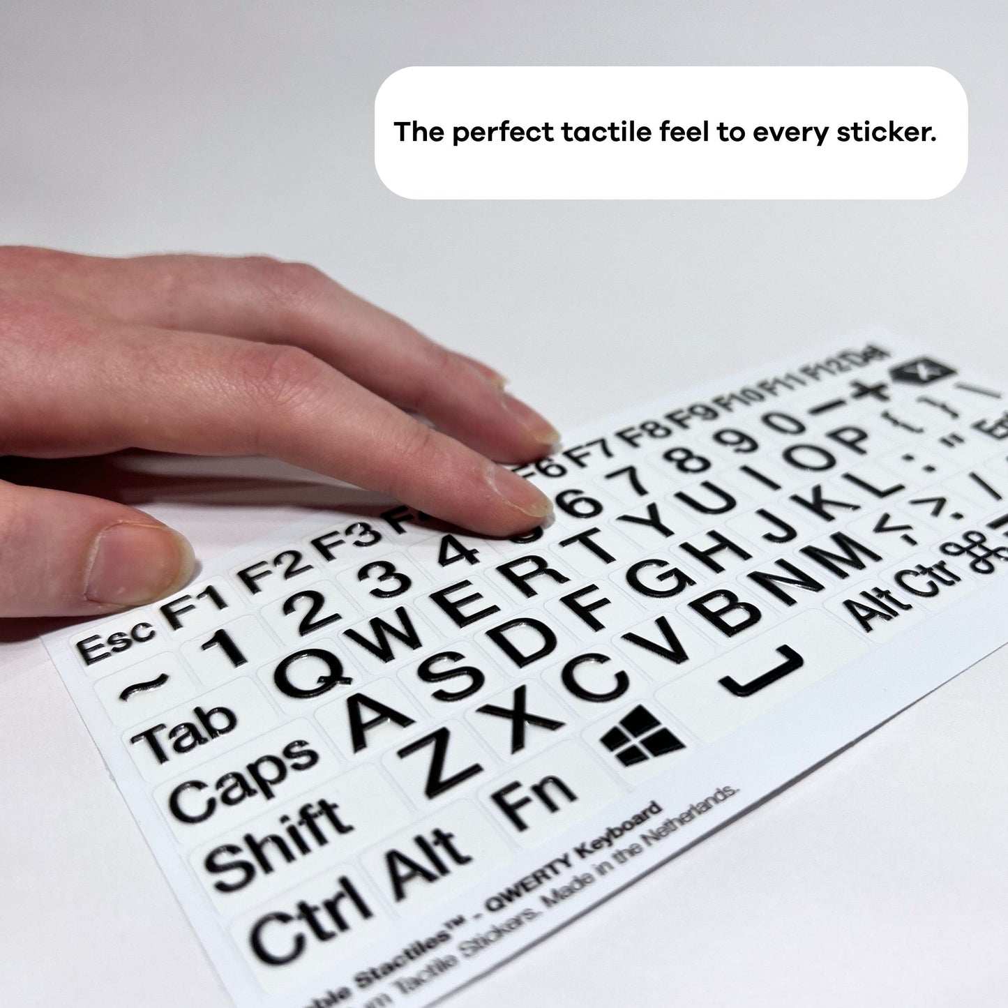 A hand moving across a sheet of Stactiles QWERTY keyboard stickers on a flat surface. At the top right in a rounded white speech-bubble shape, black text reads: “The perfect tactile feel to every sticker.”