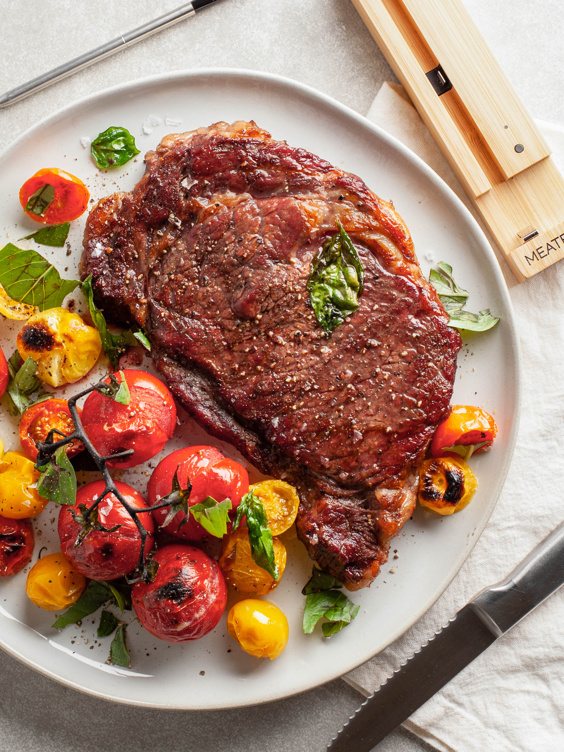 A close-up of a cooked steak with a Meater Plus thermometer, placed on a plate with sliced tomatoes on a table.
