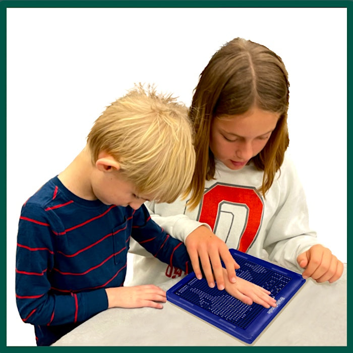 A boy with blonde hair about seven years old and wearing a blue stripe shirt, is standing with his big sister about 11 and with shoulder length, straight brown hair, in a white sweatshirt. She is guiding his hand on the BrailleDoodle to feel the braille.