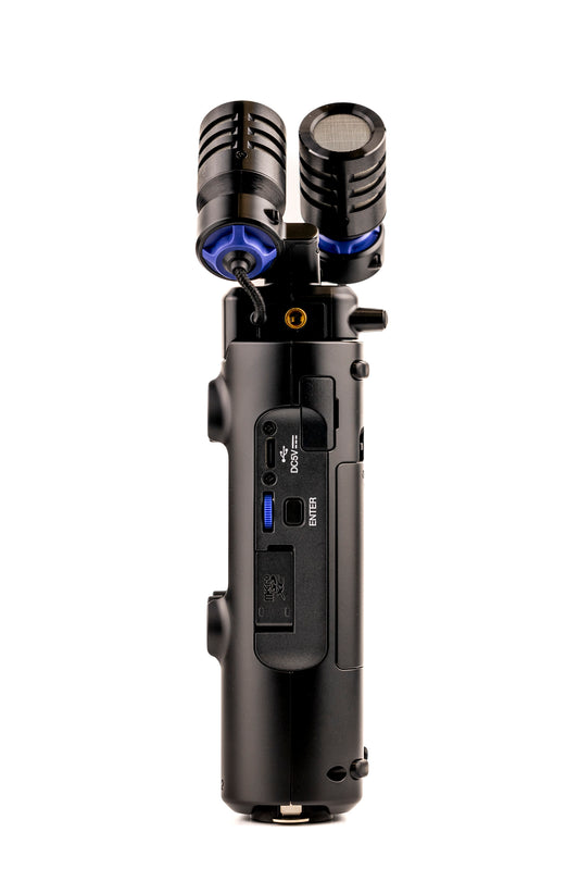 Side view of the Zoom H5studio handheld audio recorder, featuring two large X/Y stereo microphones with black grilles and blue shock mounts on top. The recorder has a matte black body with visible controls including a microSD slot, USB port, headphone jack, a blue enter dial, and a threaded mount.