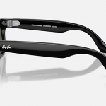 left-side view of black green Meta Ray-Bans, showing both thick curved arms from the left, with the Ray‑Ban logo on the outer temple and small white product text along the inside of the front arm, resting on a light surface.