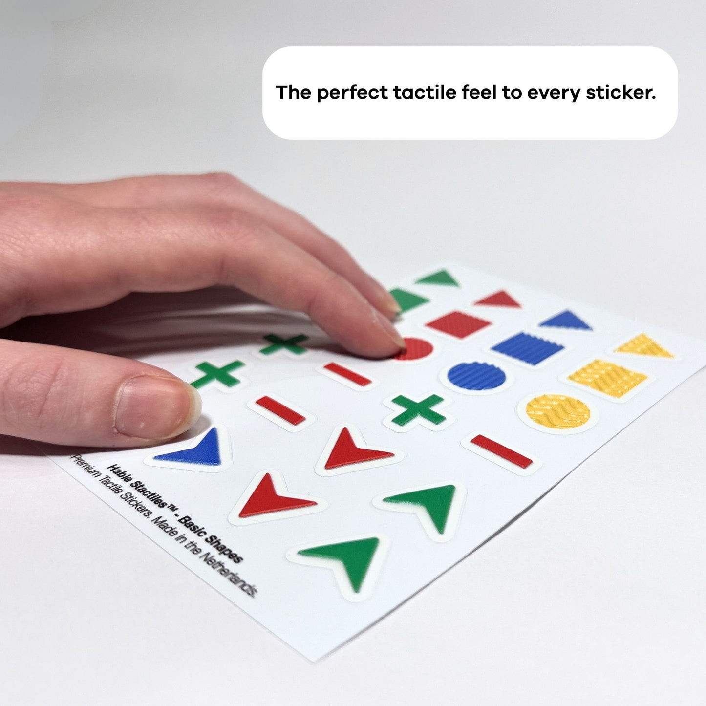 A hand moving over a sheet of Stactiles Basic Shapes tactile stickers lying on a flat surface. The white sheet has raised, colorful geometric shapes such as arrows, plus and minus signs, circles, triangles, and textured disks in red, green, blue, and yellow. At the top, inside a rounded white text bubble, it says: “The perfect tactile feel to every sticker.”