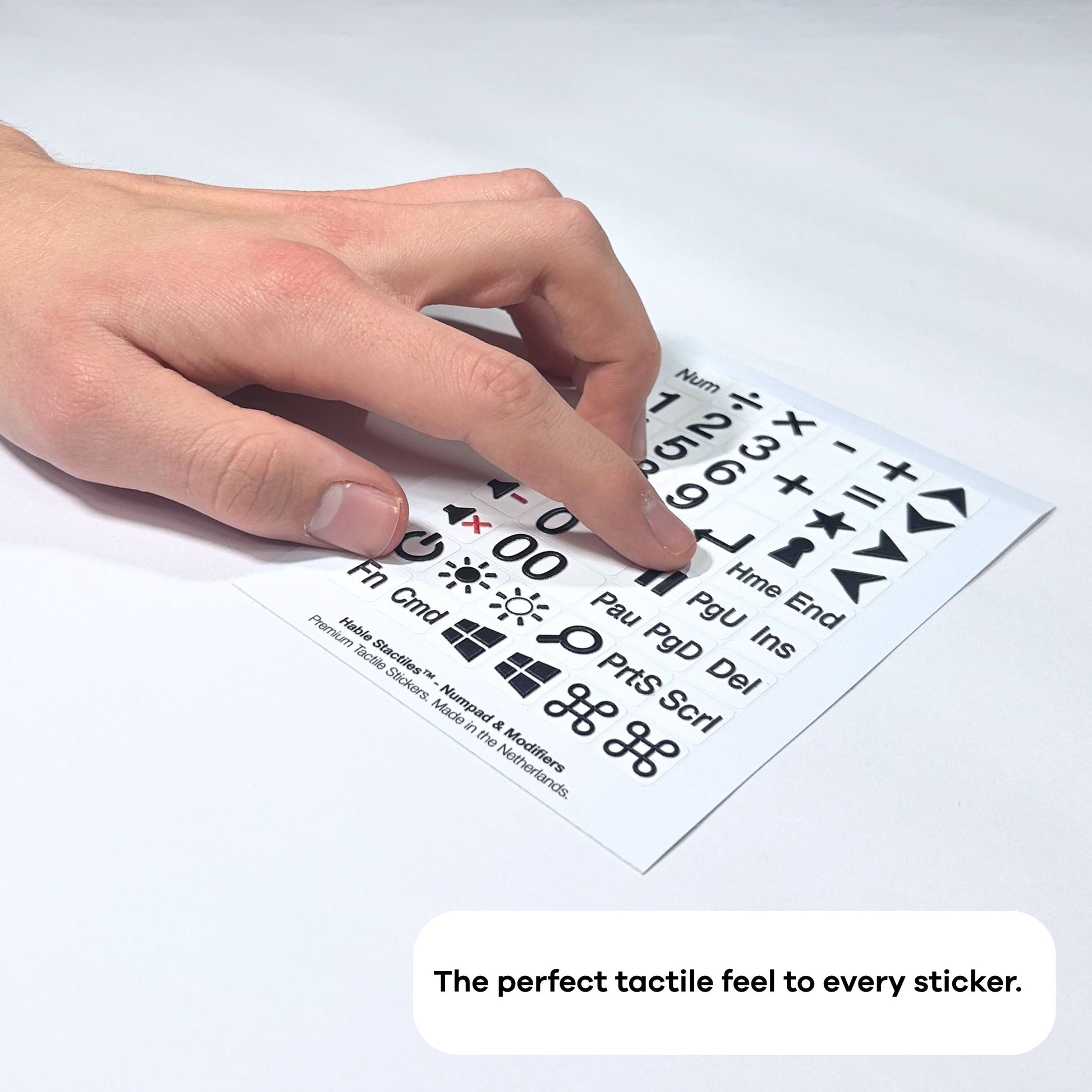 A hand moving over a sheet of Stactiles Numpad & Modifiers tactile stickers on a flat surface. The white sheet shows raised black numbers, math symbols, navigation keys like Home and End, power and brightness icons, Windows and Command symbols, and other modifiers. At the bottom of the image, in a rounded white text bubble, it says: “The perfect tactile feel to every sticker.”