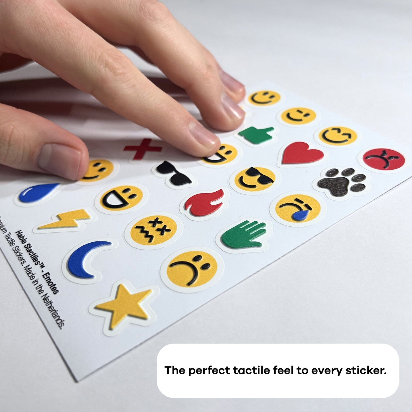 A hand moving over a sheet of Stactiles Emojis tactile stickers on a flat surface. The white sheet is filled with raised, colorful emoji-style icons: yellow faces with different expressions, hearts, flames, a paw print, a lightning bolt, a crescent moon, a star, and other symbols. At the bottom of the image, inside a rounded white text bubble, it reads: “The perfect tactile feel to every sticker.”