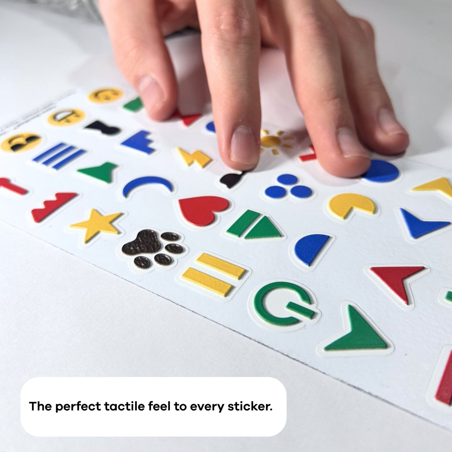 A hand moving across a sheet of Stactiles Icons stickers on a flat surface. The sheet includes raised shapes such as arrows, hearts, stars, circles, crescents, a paw print, and other geometric icons in bright colors like red, yellow, blue, green, and black. At the bottom of the image, inside a rounded white text bubble, it reads: “The perfect tactile feel to every sticker.”