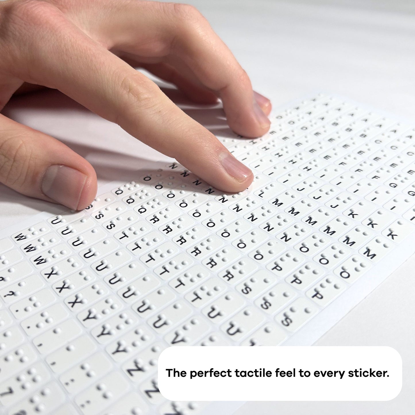 A person uses both hands to feel a Stactiles letters sheet, exploring the raised alphabet stickers arranged in a grid. Another sheet with tactile icons is partially visible beside it on the table.