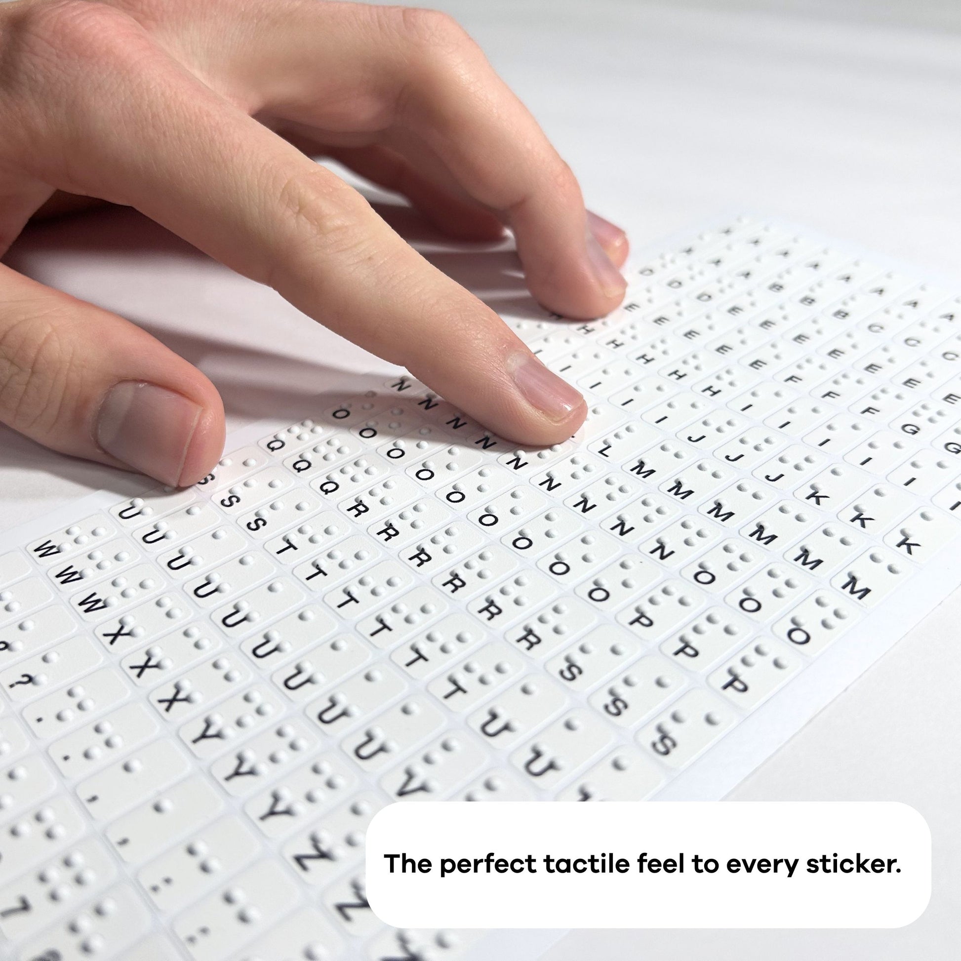A person uses both hands to feel a Stactiles letters sheet, exploring the raised alphabet stickers arranged in a grid. Another sheet with tactile icons is partially visible beside it on the table.