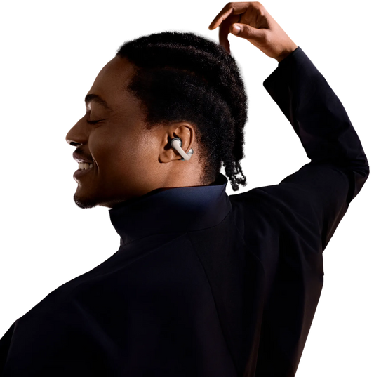 A smiling man with short dreadlocks and eyes closed, his head slightly turned to the side, wearing a dark high-collar jacket. He sports a Shokz OpenDots1 earbud in one ear. One arm is raised, with his hand positioned above his head in a graceful pose.